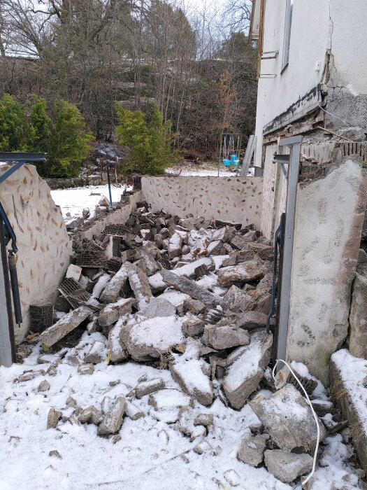 Rasat betongtak vid husfasad, omgiven av snö och bråte, med nerfallna rester av väggbeklädnad. Omgivning av träd och buskar i bakgrunden.