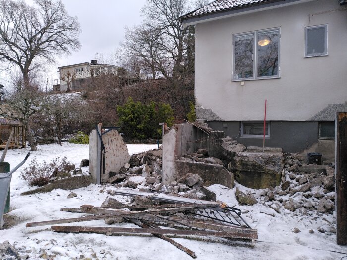 Rivning av betongkonstruktion vid ett hus, med synliga rester av tak och väggar på marken, i en snötäckt trädgård.