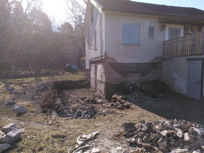 Riven tomt med rester av tak och väggar, betongbitar spridda på gräset framför ett hus, beläget vid en skog.