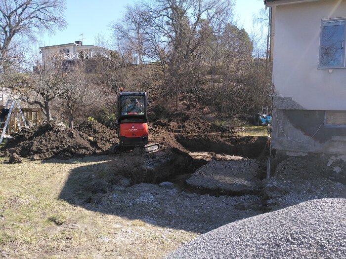 Grävmaskin gräver en grop nära ett hus för en ny källare, omgiven av jordhögar och grus.