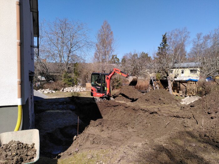 Grävmaskin som gräver en grund för en källare vid ett hus, med jordhögar och träd i bakgrunden. Underlag med markduk och makadam syns.