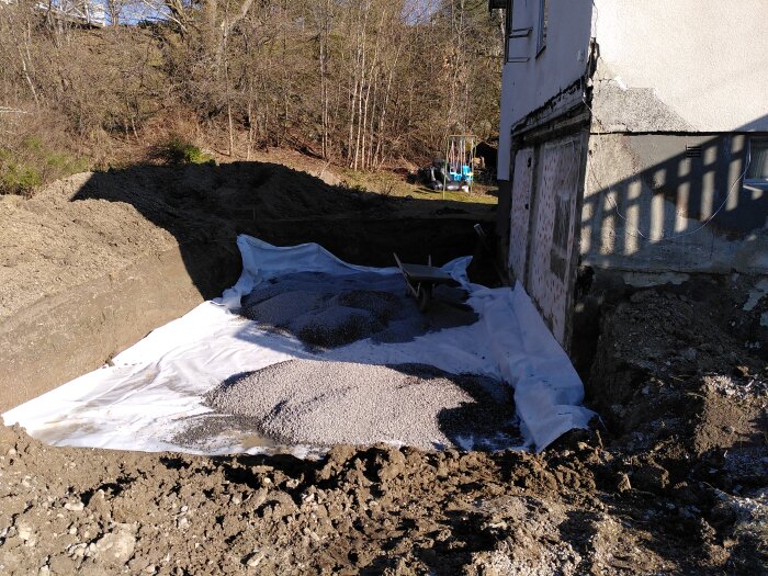 Utgrävt område vid husvägg med markduk och makadam, förberett för ny källare. Skottkärra synlig i bakgrunden.