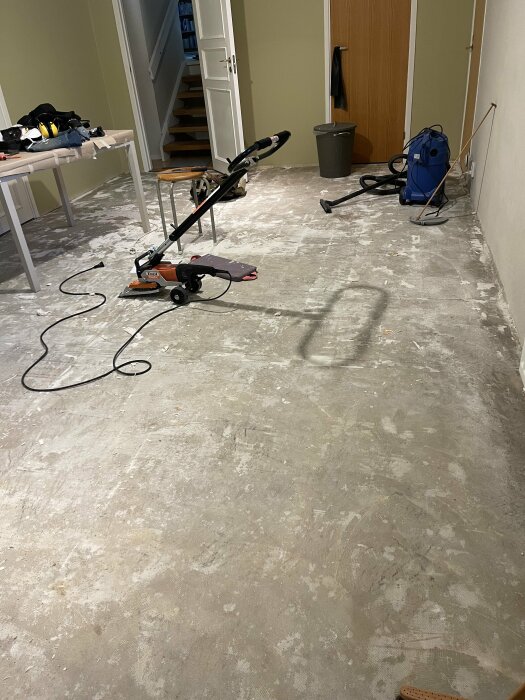 Betonggolv med limrester i källare, utrustning för borttagning av plastmatta syns i bilden, inklusive en mattstripper.