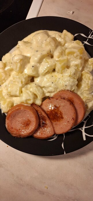 Stuvad potatis och stekt falukorv på en svart tallrik.