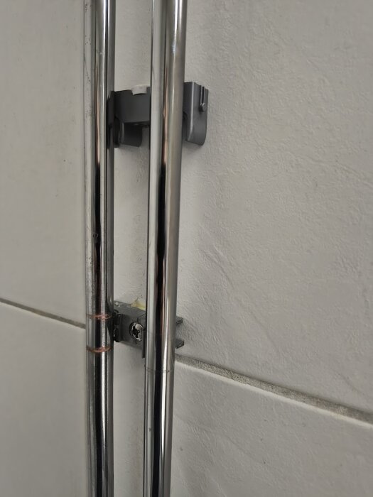 En rörhållare i duschen med spruckna delar, monterad på väggkakel med två vertikala kromrör som delvis döljer hållaren.