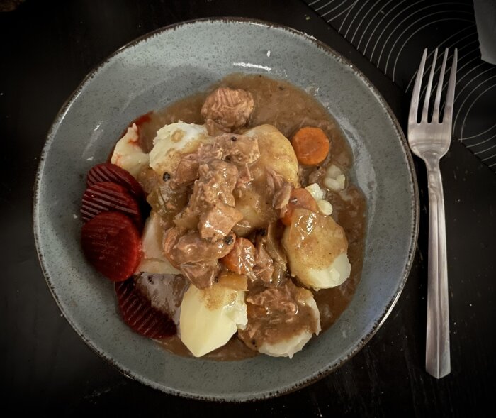Kalops med kött, kokt potatis, morötter och inlagda rödbetor på en blå tallrik.