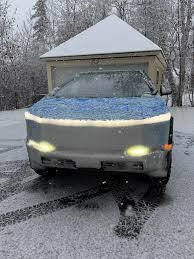 Cybertruck med snö på fronten framför helljusen, parkerad på en snöig uppfart framför ett garage.