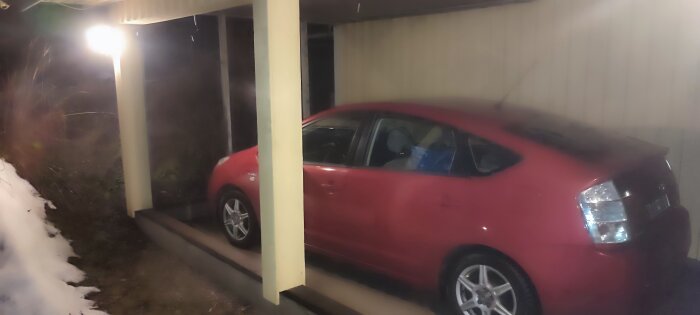 Röd bil under carport vid garage med snö och betongkant, nattbild.