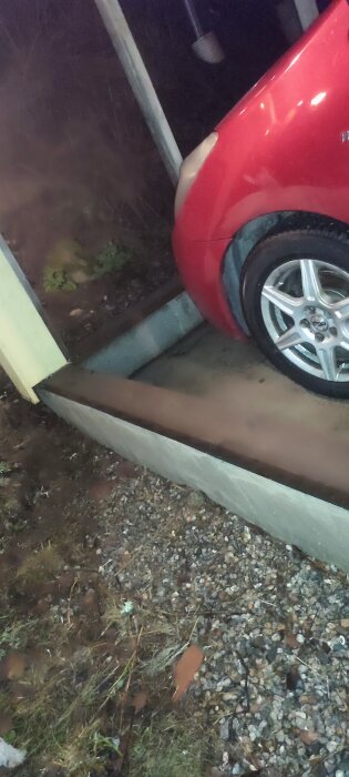 Röd bil i en carport med betongplatta och upphöjd kant av lättbetong, omgiven av stenig mark och växtlighet.