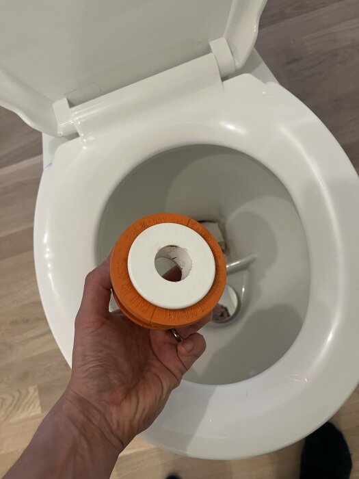 En hand håller en orange och vit del ovanför en öppen vit toalettstol av märket Wostman Ecoflush.