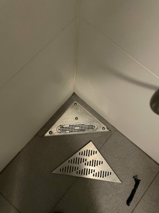 Hörn av ett badrumsgolv med en triangel formad Purus Corner hörnbrunn och en borttagen brunnslock i rostfritt stål på grå klinker.