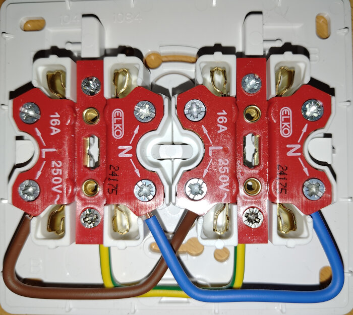Eluttag med parallella kopplingsplattor, märkta 16A 250V; kablar i brunt, blått och grönt monterade i terminalerna на bakstycket.