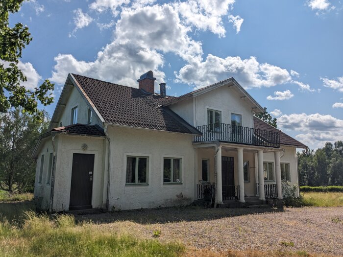 Vit tvåvånings villa med balkong och tegeltak, belägen på landet under en solig, delvis molnig himmel. Grusad uppfart och omgiven av vegetation.