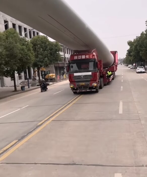 Lastbil transporterar stor turbinbladsliknande föremål genom stadsgata med byggnader och träd.