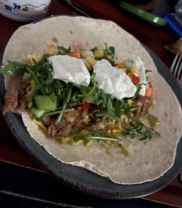 Pulled pork i mjuk tortilla med sallad, picklad rödlök, guacamole, riven ost, inlagd chili, salsa och vitlökssås på en tallrik.