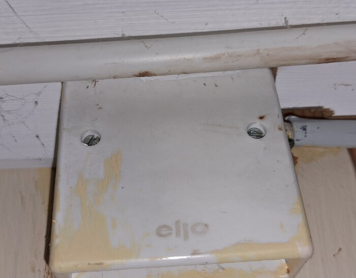 Gammalt taklampauttag från 60-talet med texten "ello" och sladdar, monterat på en sliten yta med synliga skruvar.