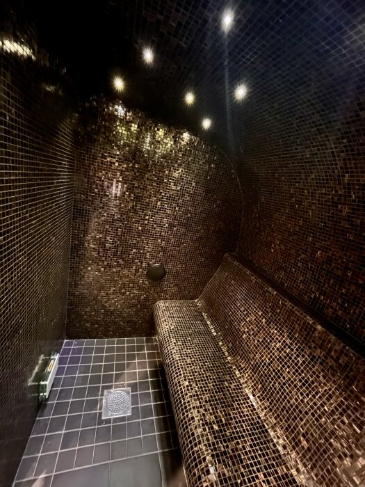 Inne i en ångbastu med mosaikväggar och belysning i taket.