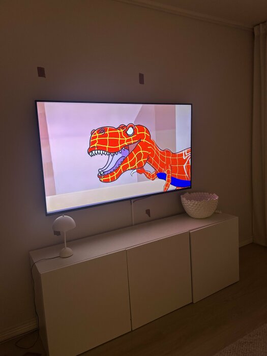 Vägghängd TV med tecknad dinosaurie i vardagsrum, tejpbitar på väggen markerar möjligt förberett dörröppning.