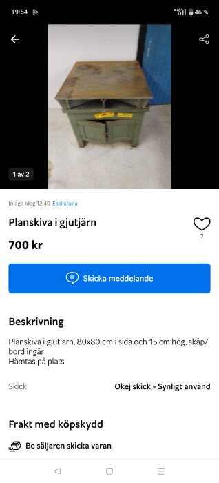 Gjutjärnsskiva på grön metallmöbel med dörrar, används som bord. Skivan är sliten och ytan har rostfläckar. Fotad mot en vit vägg och blå dörr.