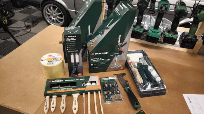 Olika verktyg och tillbehör från Lidl, inklusive Parkside-verktyg, penslar, maskeringstejp och borrbits, placerade på ett bord i ett garage.