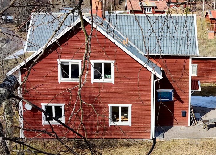 Rött hus med grått tak och vita fönster. Väggarna är täckta med horisontella paneler, omgivna av trädgrenar, utan dekorativa midjor.