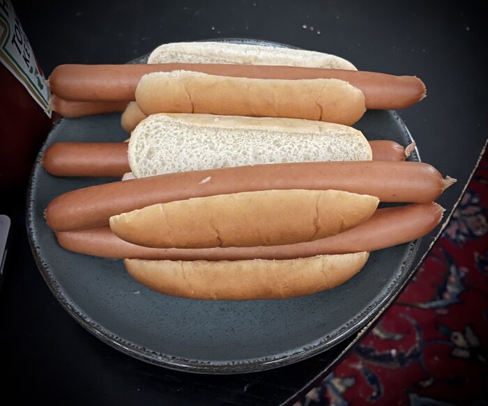 Tallrik med fem varmkorvar i bröd på ett mörkt bord, bredvid en flaska ketchup.
