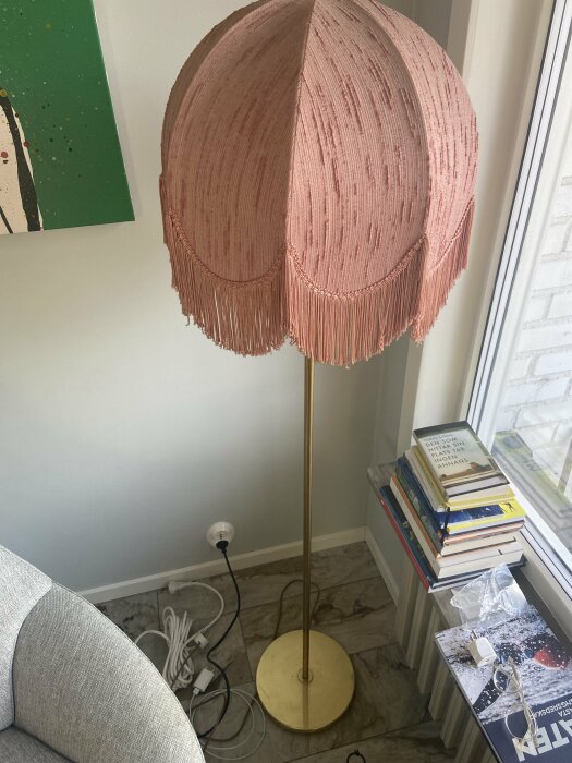 En rosa second hand-lampa med fransig lampskärm står bredvid en bokhög vid ett fönster. Sladdar syns på golvet, och lampan är ansluten i ett uttag.