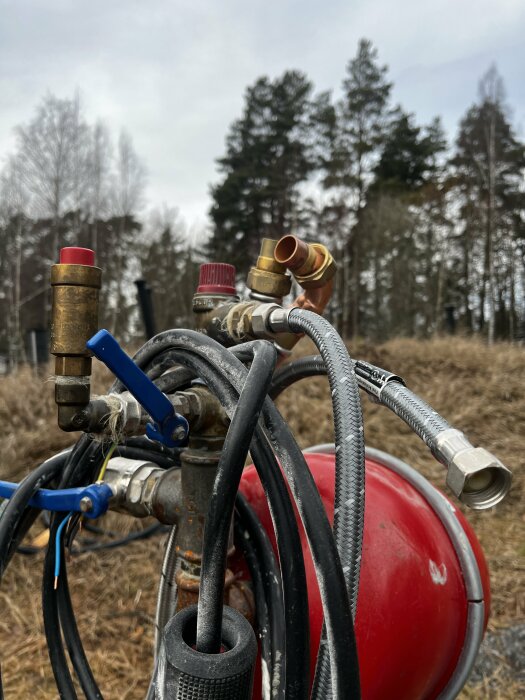 Röd byggpanna med slangar och ventiler utomhus, omgiven av träd och gräs.