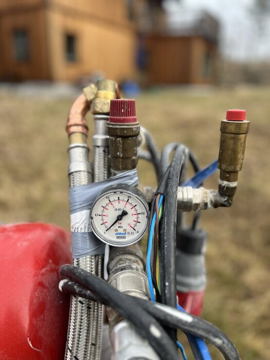 Byggpanna 9kW med tryckmätare och ledningar i förgrunden; rött träbyggnad suddigt i bakgrunden.