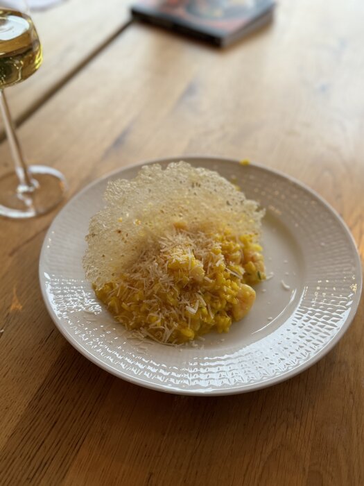 Saffransrisotto med vannameiräkor och citronflarn på vitt fat; ett glas vin syns bredvid på ett träbord.