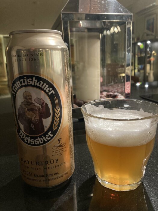 En ölburk med Franziskaner Weissbier och ett glas öl med skum ovanpå, placerade på en mörk yta.