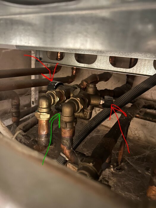Blandningsventiler till gaspanna med två rattar, en med grön pil och en med röd pil. Bakom ses kopparrör och en metallplatta.