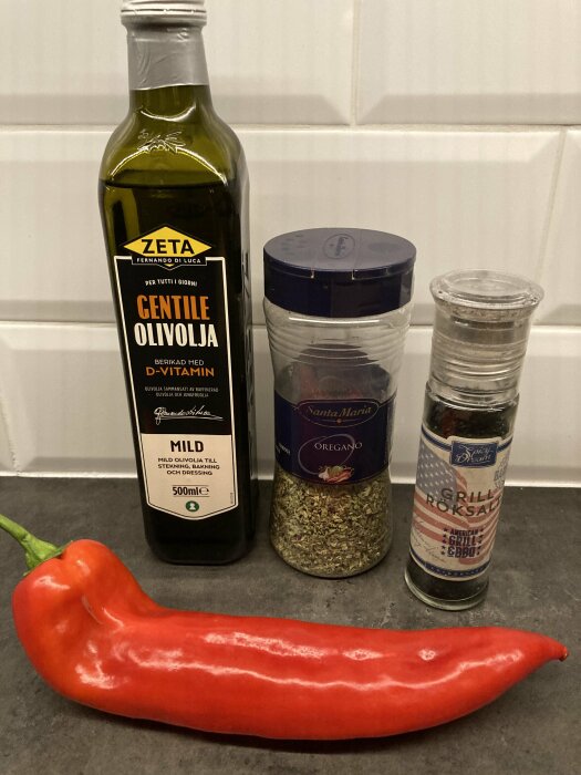 Flaska olivolja, burk med oregano, flaska grillsalt och en röd paprika på en mörk bänkskiva.