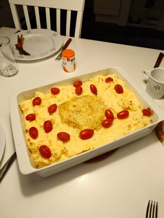 Planksteksgratäng med potatismos och tomater i en ugnsform, serverad på ett bord bredvid en tallrik med sparris och bacon.