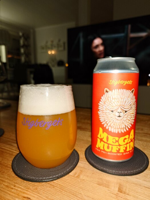 Ölglas med skummande Stigbergets öl bredvid en Mega Muffin burk på ett bord.