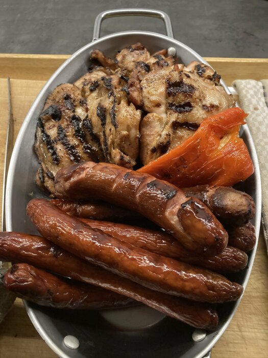 Grillad paprika, korvar och kött i en metallbricka på träskiva.