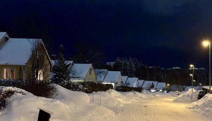 Snötäckt gatvy med radhus i vinterlandskap på kvällen, gatubelysning och snödrivor längs vägen.