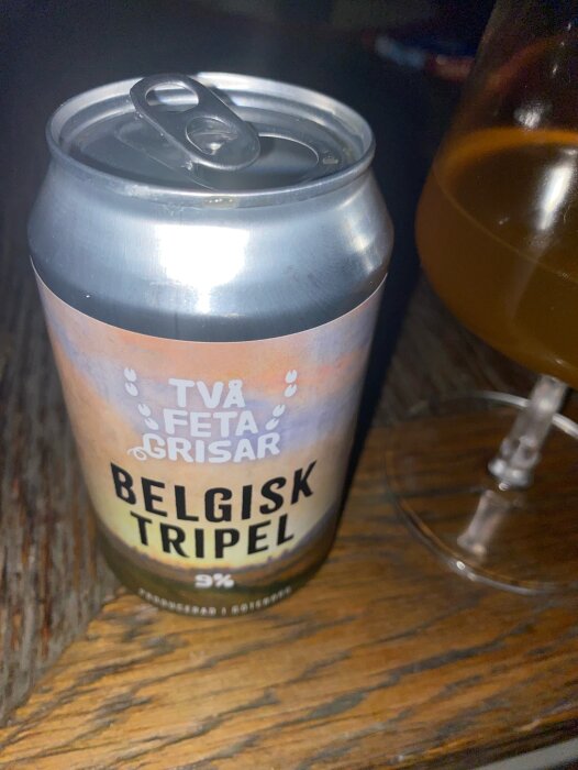 En ölburk märkt "Två Feta Grisar Belgisk Tripel" bredvid ett glas med samma öl på ett träbord.