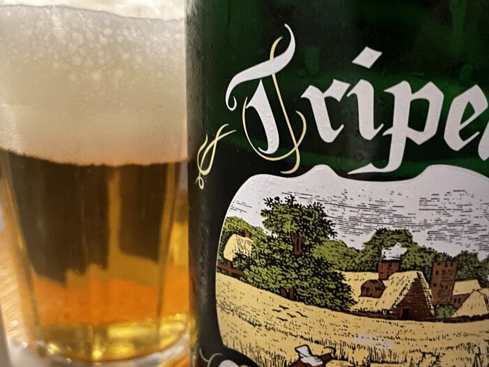 Stor flaska med en etikett som visar en landskapsbild med hus och en äng. Ett glas med skummande öl syns bredvid.
