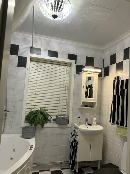 Modernt badrum med vitt och svart kakel, jacuzzi, takdusch och belysning frÄn Plejd. VÀxt vid fönstret och handfat med spegelskÄp.