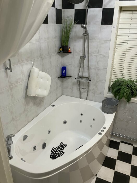 Hörnbadkar med massagefunktion i svartvitt kaklat badrum; ovanför syns en takdusch, vÀxt, och fönster med persienner.