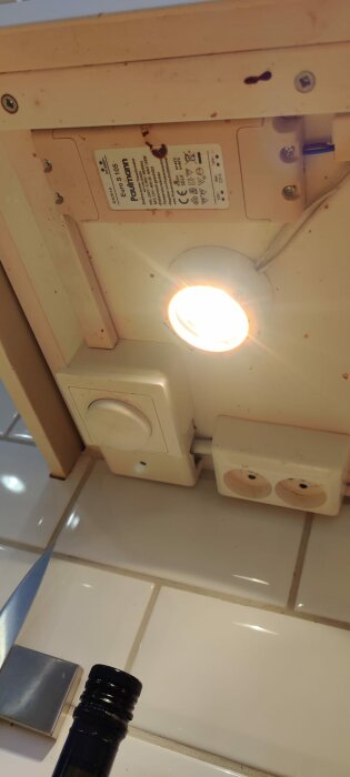 Dimmer, strömuttag och en takspotlight syns tillsammans på en köksvägg med en flaska i förgrunden.