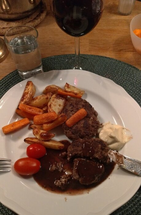 Tallrik med köttfärsbiffar, ugnsrostade grönsaker, två körsbärstomater, sås och bearnaisesås. Ett glas rött vin och vatten i bakgrunden.