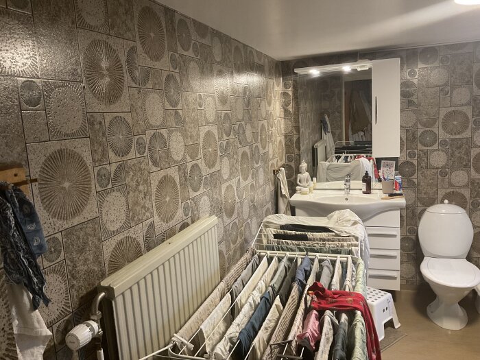 Badrum med mönstrade väggkakelplattor, vit toalett och handfat med spegel. Klädställning med torkande kläder framför elementen.