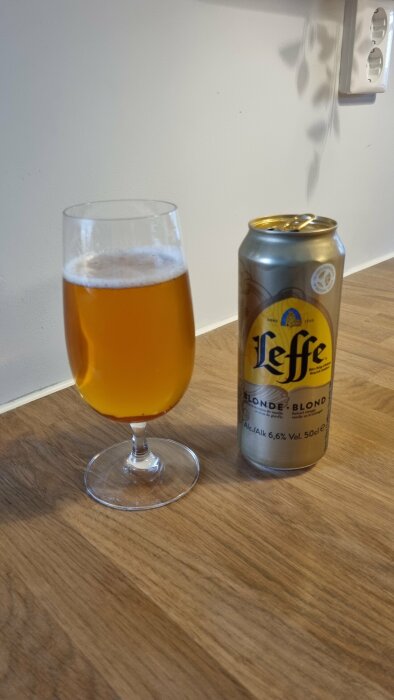 Ett glas med gyllene öl bredvid en burk Leffe Blond på ett träbord.