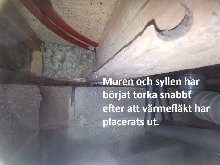 Ingång till krypgrund med murad betongvägg och syll, isolering och rör. Text: "Muren och syllen har börjat torka snabbt efter att värmefläkt har placerats ut.