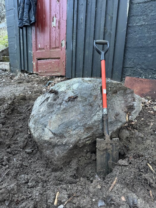 Stor sten vid röd dörr med spade lutad mot stenen i en jordig omgivning.