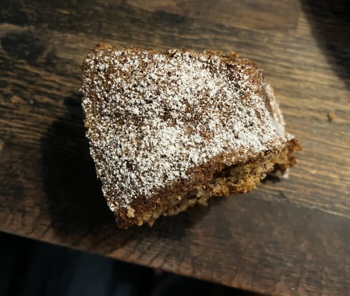 En bit banankaka med florsocker på toppen ligger på ett mörkt träbord.