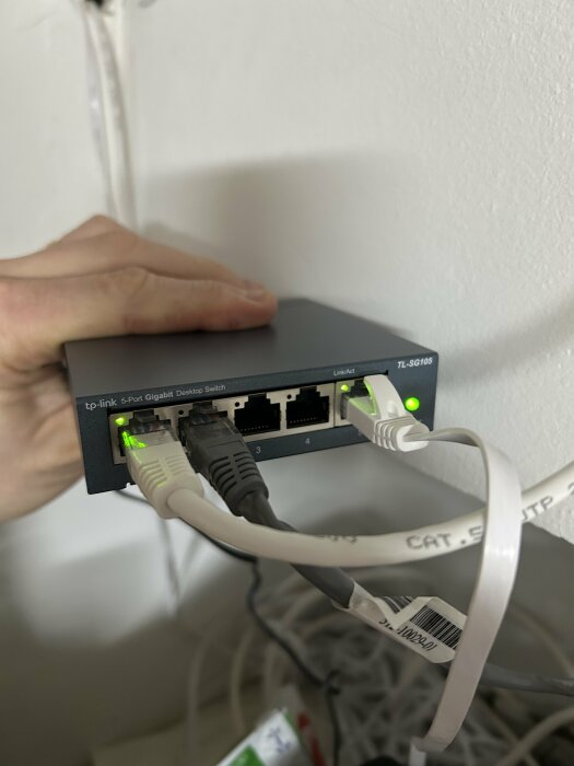 En hand håller i en TP-Link 5-Port Gigabit Desktop Switch med flera nätverkskablar anslutna; en kabel längst till höger kommer från routern.