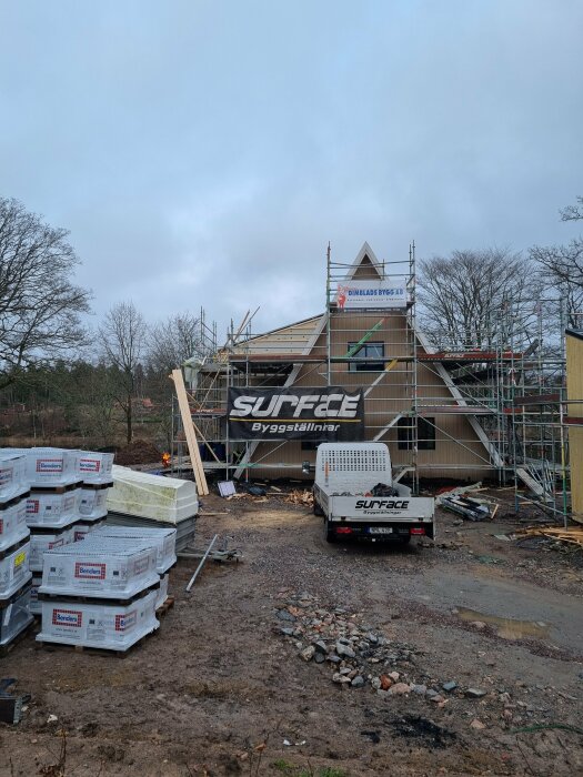 Hus under renovering med byggnadsställningar och lastbil från byggfirma framför. Marken är lerig och byggmaterial ligger staplat på sidan.